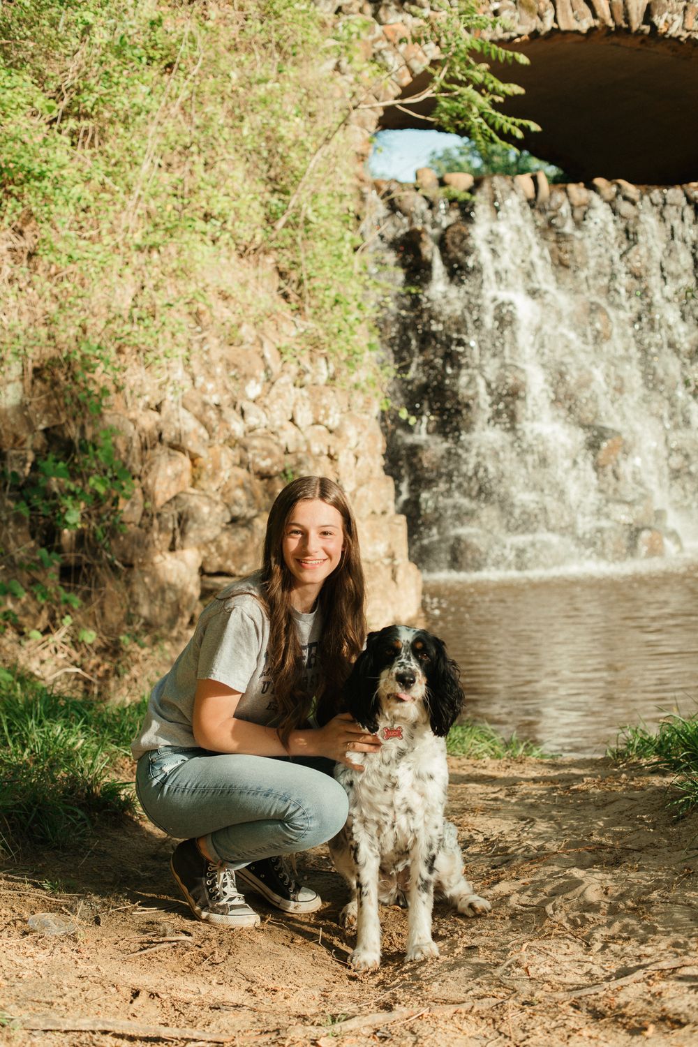 A black and white dog sitting with someone by a small waterfall in a natural setting.