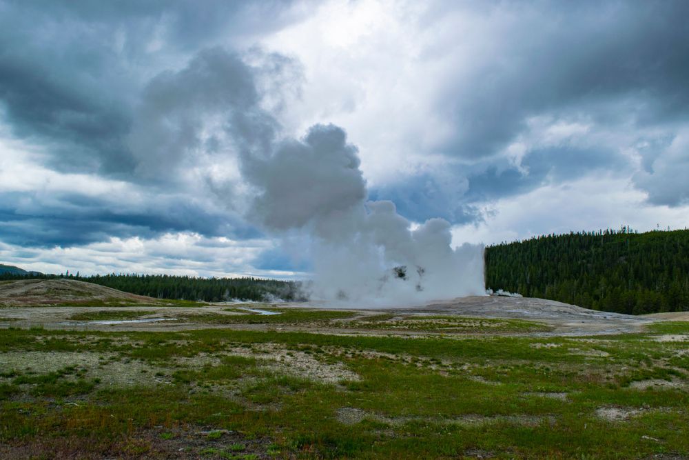Steam rises from a geyser in Yellowstone's vast landscape under dramatic storm clouds.