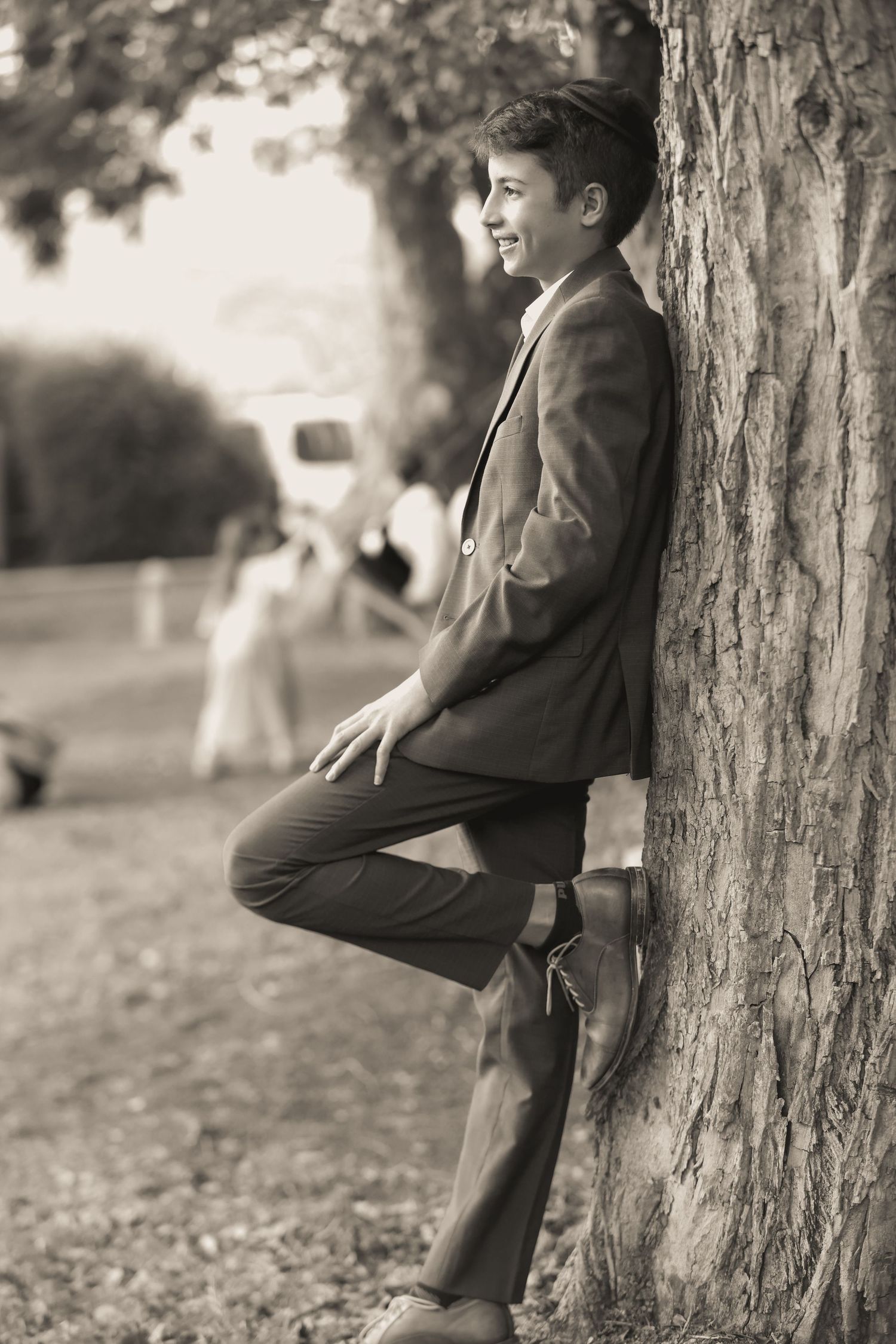 Teen boy in blazer leaning against tree trunk in sepia-toned outdoor portrait.