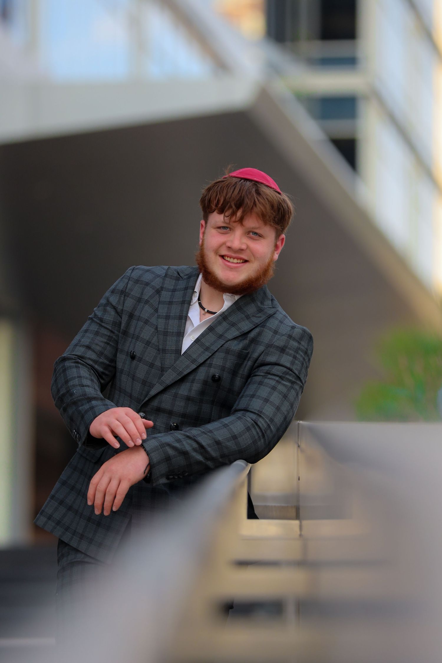 Smiling young man in plaid suit and red kippah leaning on railing at outdoor venue.