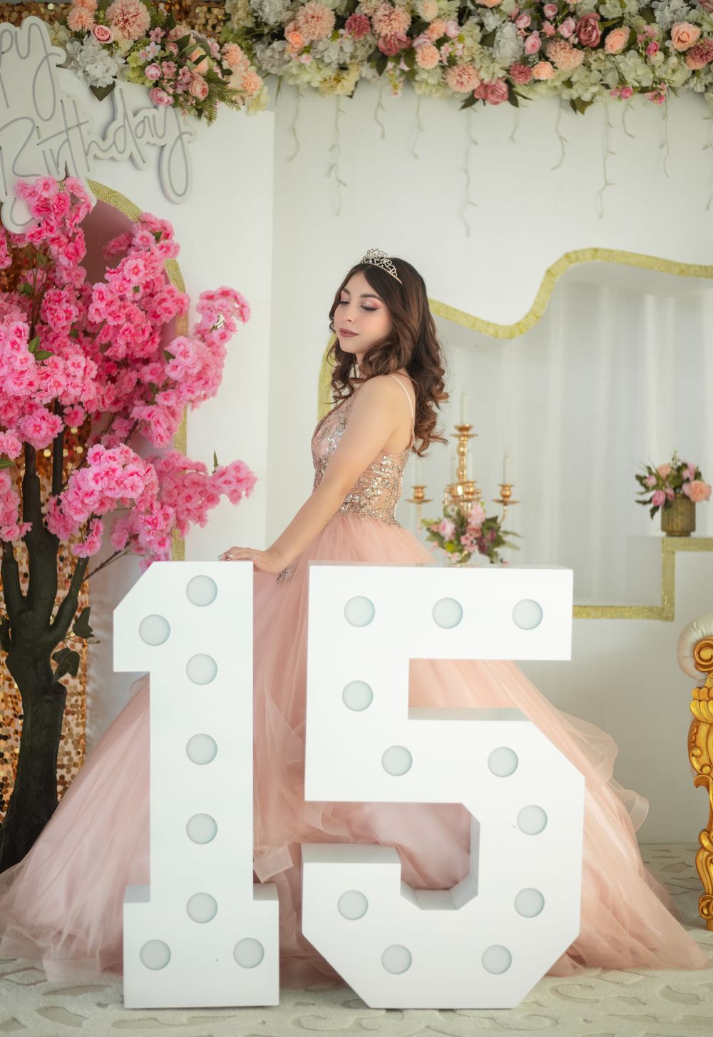 Elegant ball gown with illuminated number 15 display surrounded by pink floral arrangements and white backdrop.