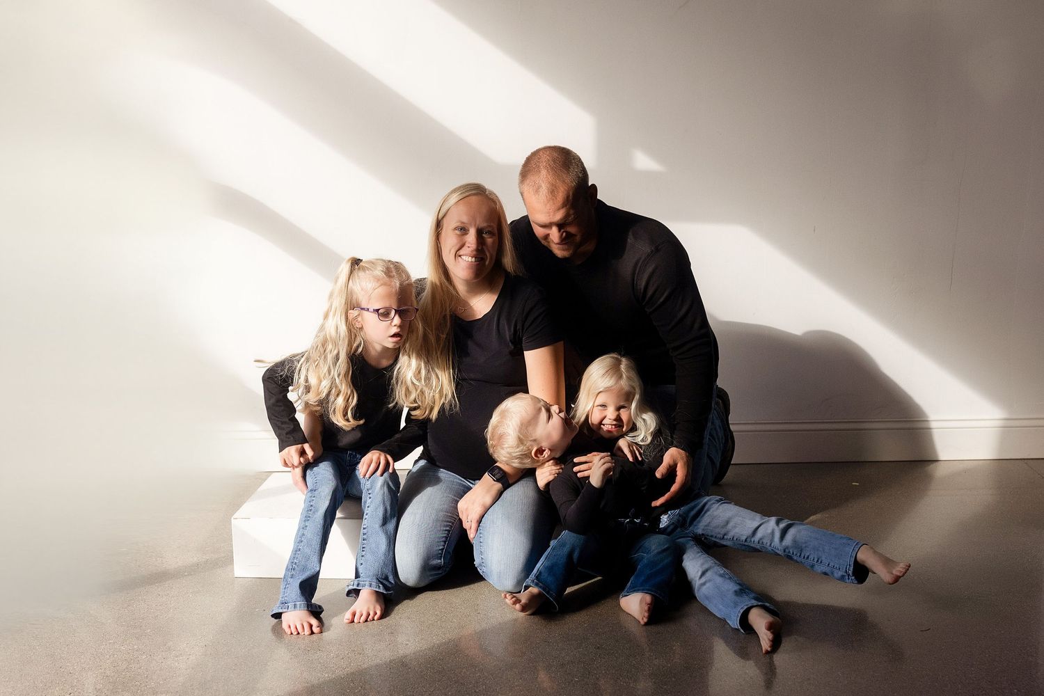 A family in casual denim sits together on a white floor in a minimalist setting with dramatic lighting and shadows.
