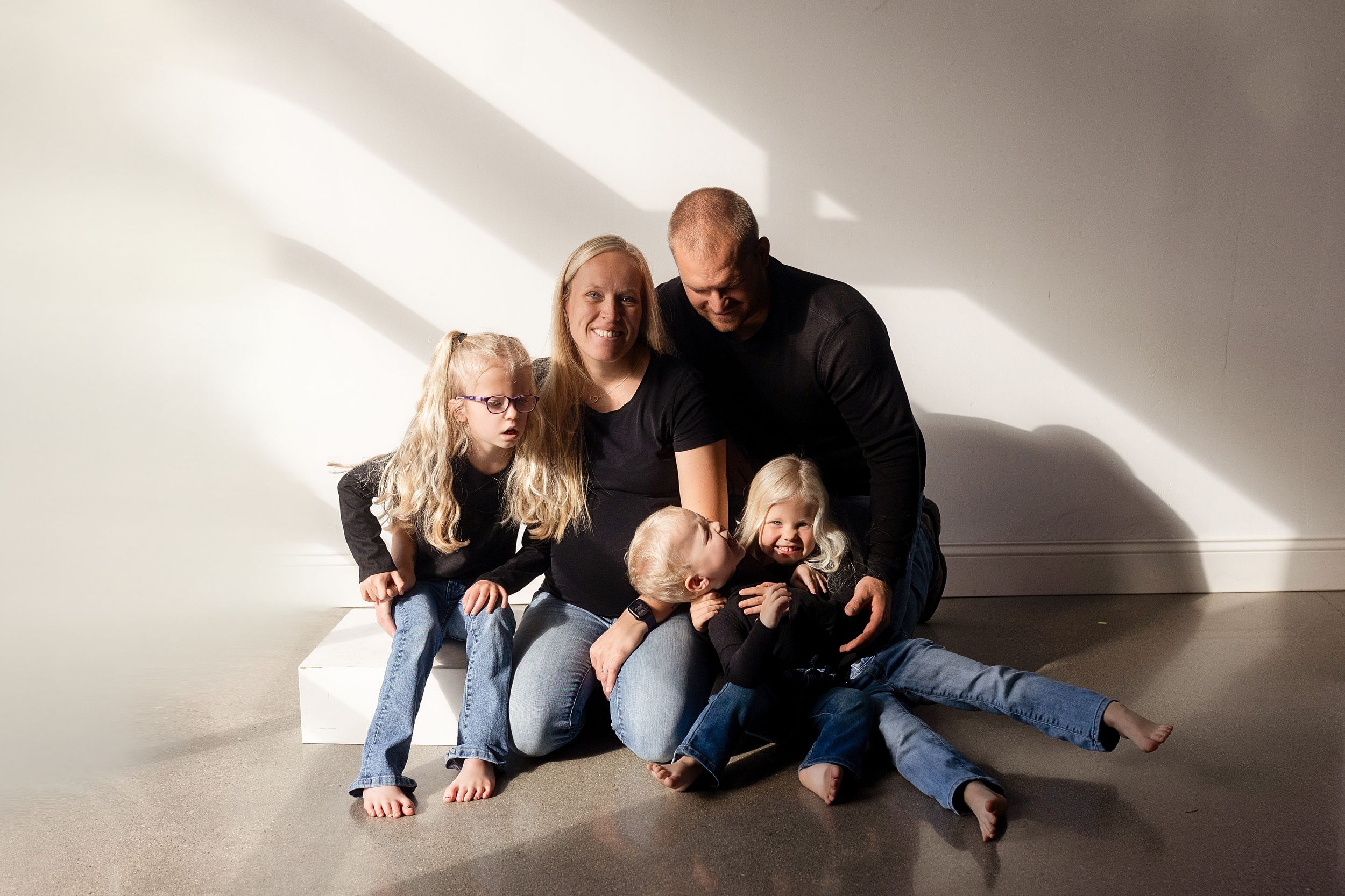 A family in casual denim sits together on a white floor in a minimalist setting with dramatic lighting and shadows.