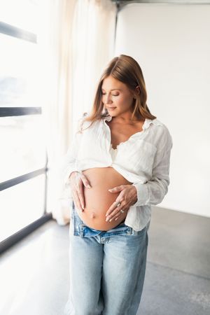 A pregnant woman in a white shirt and jeans stands by a window, gently cradling her baby bump.