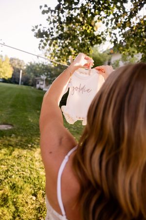 Someone holds up a small baby onesie in golden sunlight against a green lawn background.