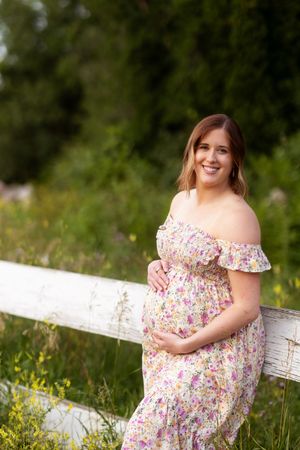 A person in a floral off-shoulder dress poses outdoors near a white fence surrounded by wildflowers in a sunny meadow.