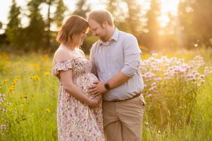 A couple embraces in a golden wildflower meadow at sunset while soft light filters through tall trees in the background.