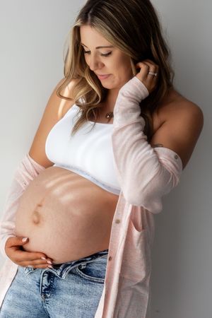 A series of maternity photos showing a pregnant woman in jeans and white top touching her belly against a gray backdrop.