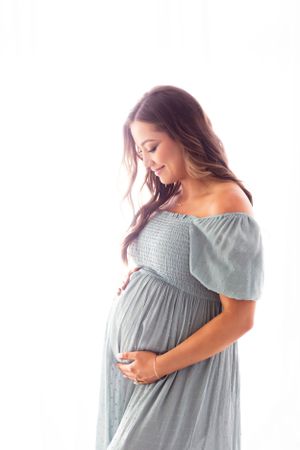 Side profile of pregnant woman in grey off-shoulder dress against white background.