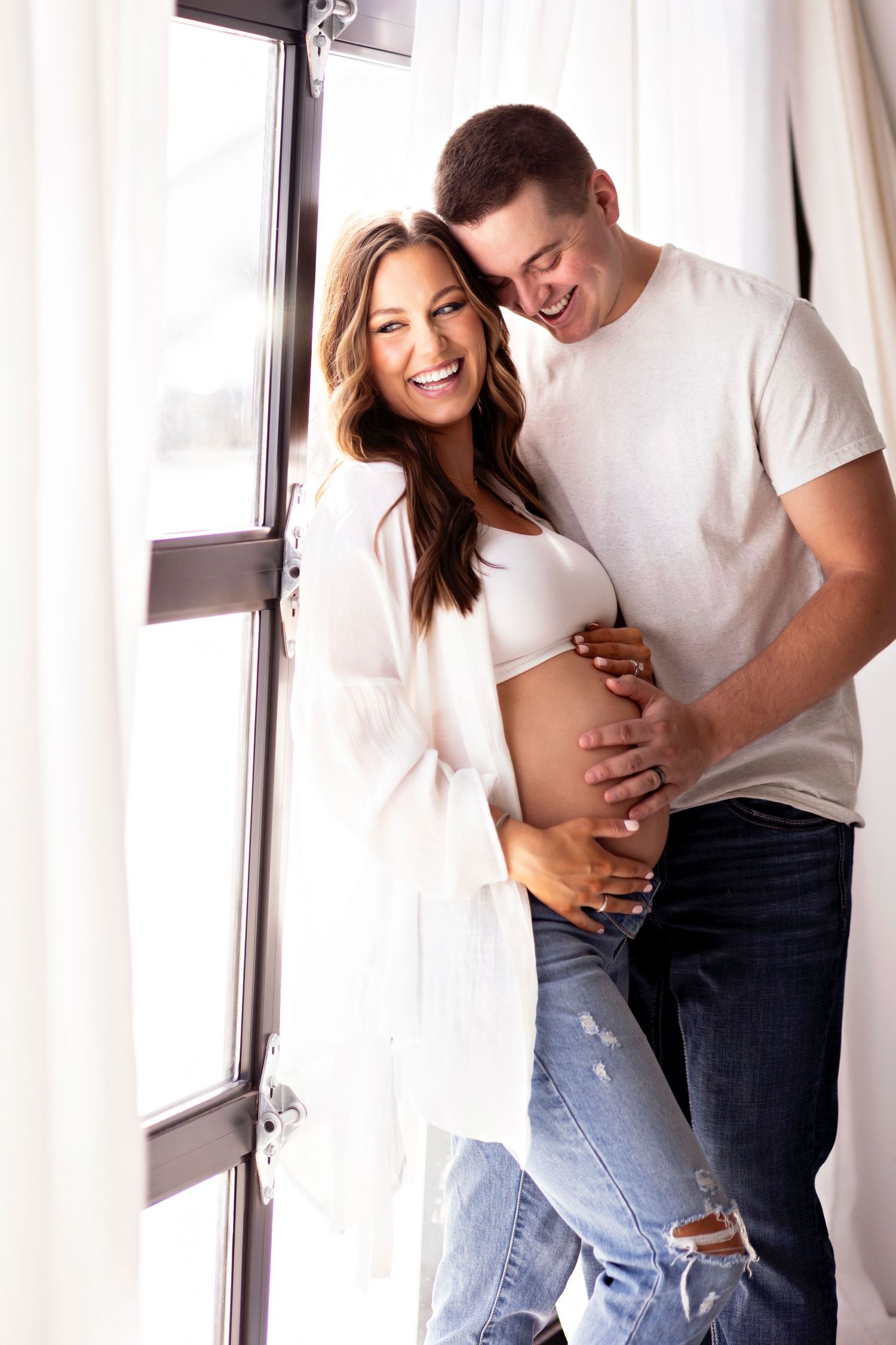 A couple shares an intimate moment by a window while embracing a pregnant belly.
