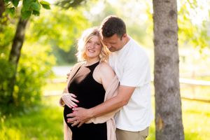 A couple embraces outdoors in a sunlit park setting with trees and a wooden fence in the background.
