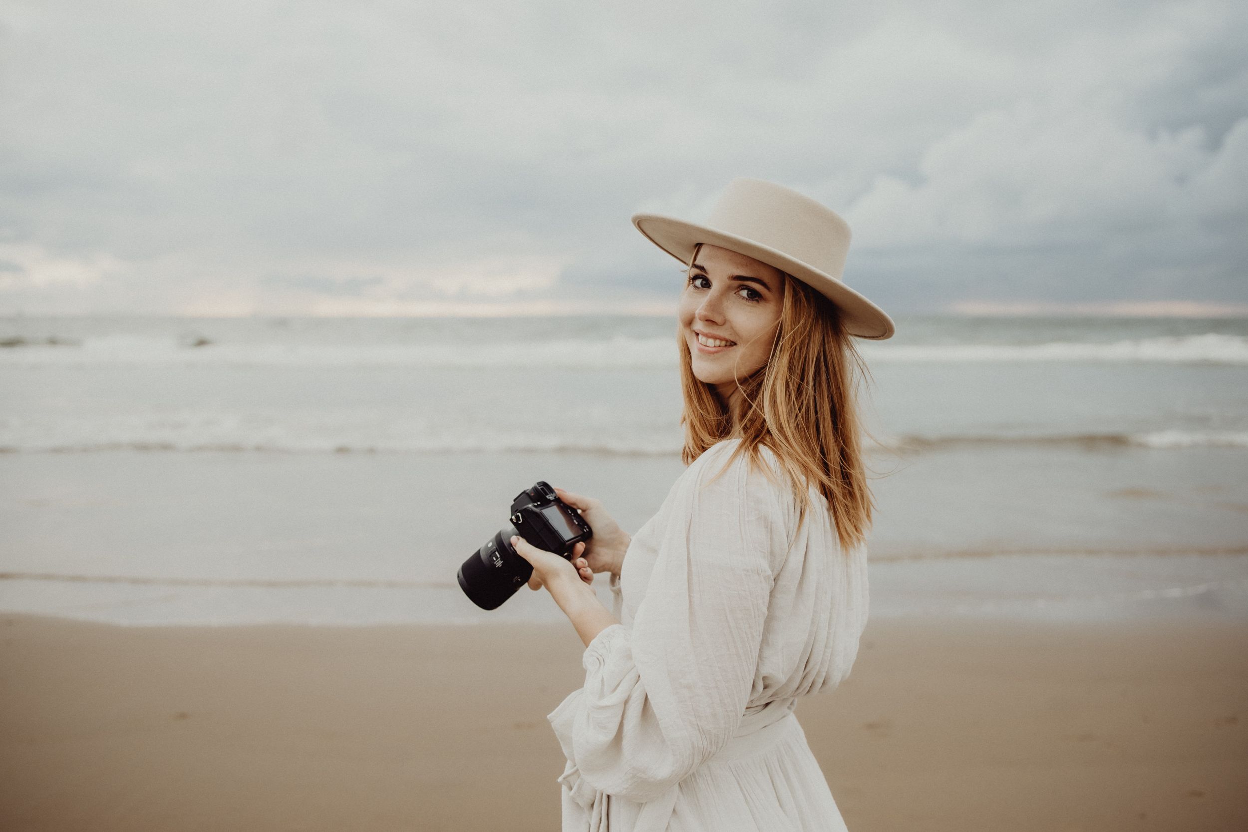 Photographer in white dress and beige hat holding camera on cloudy beach at sunset.