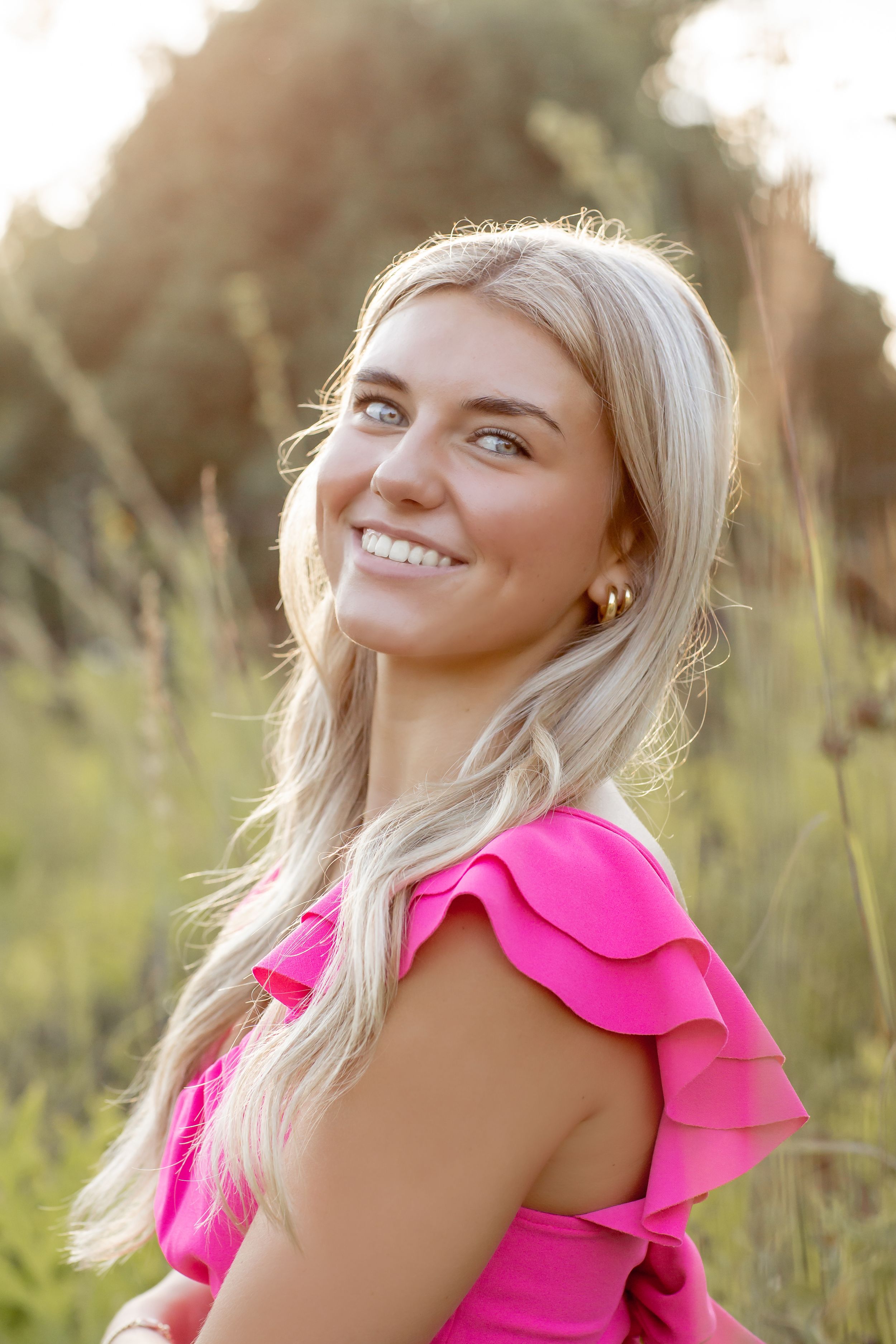 Smiling blonde woman in hot pink ruffled dress posing in sunlit field.