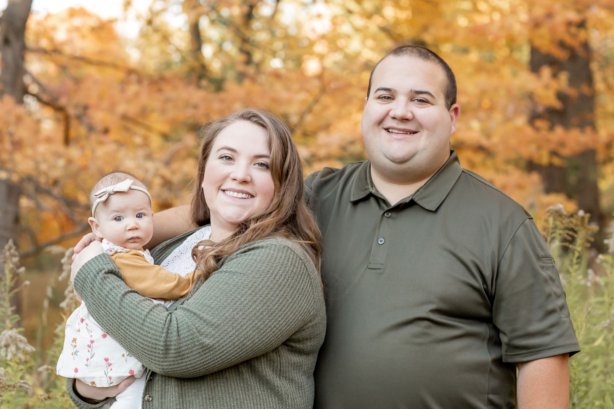 Smiling couple holding their baby girl outdoors with vibrant autumn trees in the background.