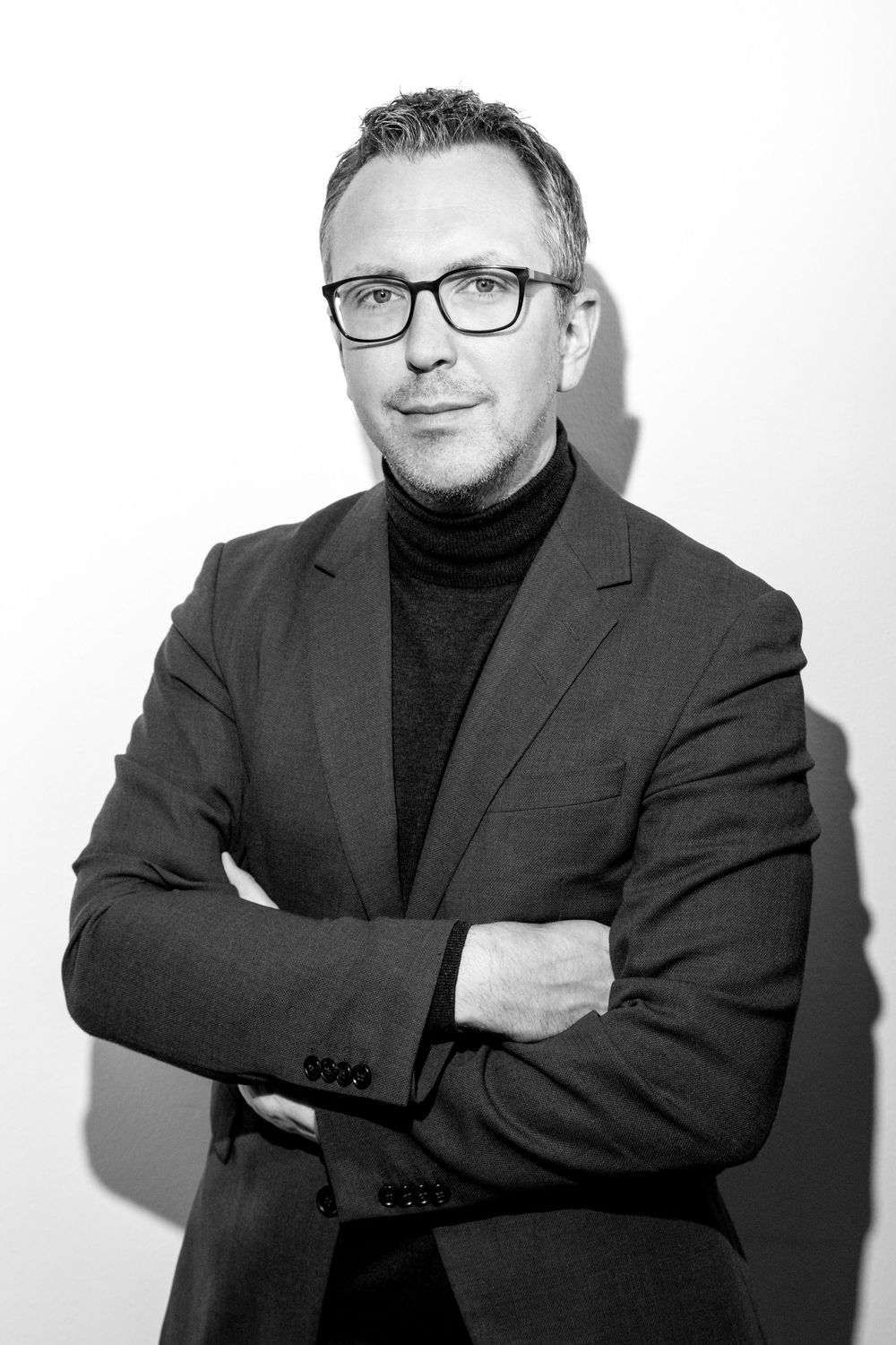 Black and white portrait series of a person wearing glasses and dark turtleneck with blazer against white background.
