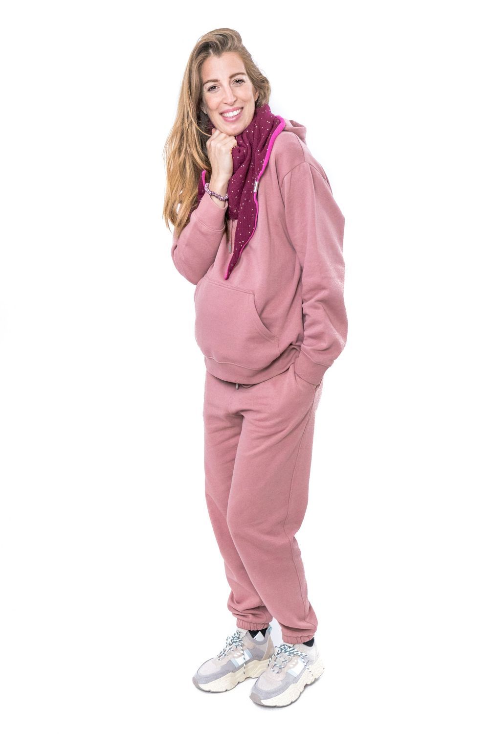 Person wearing a pink tracksuit with white sneakers poses against white background while smiling.