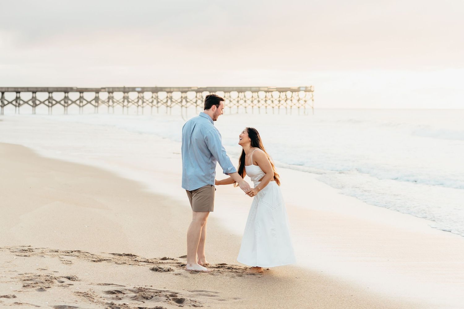 A couple stands together on a peaceful beach with a wooden pier in the background. Wilmington NC. Wrightsville beach.