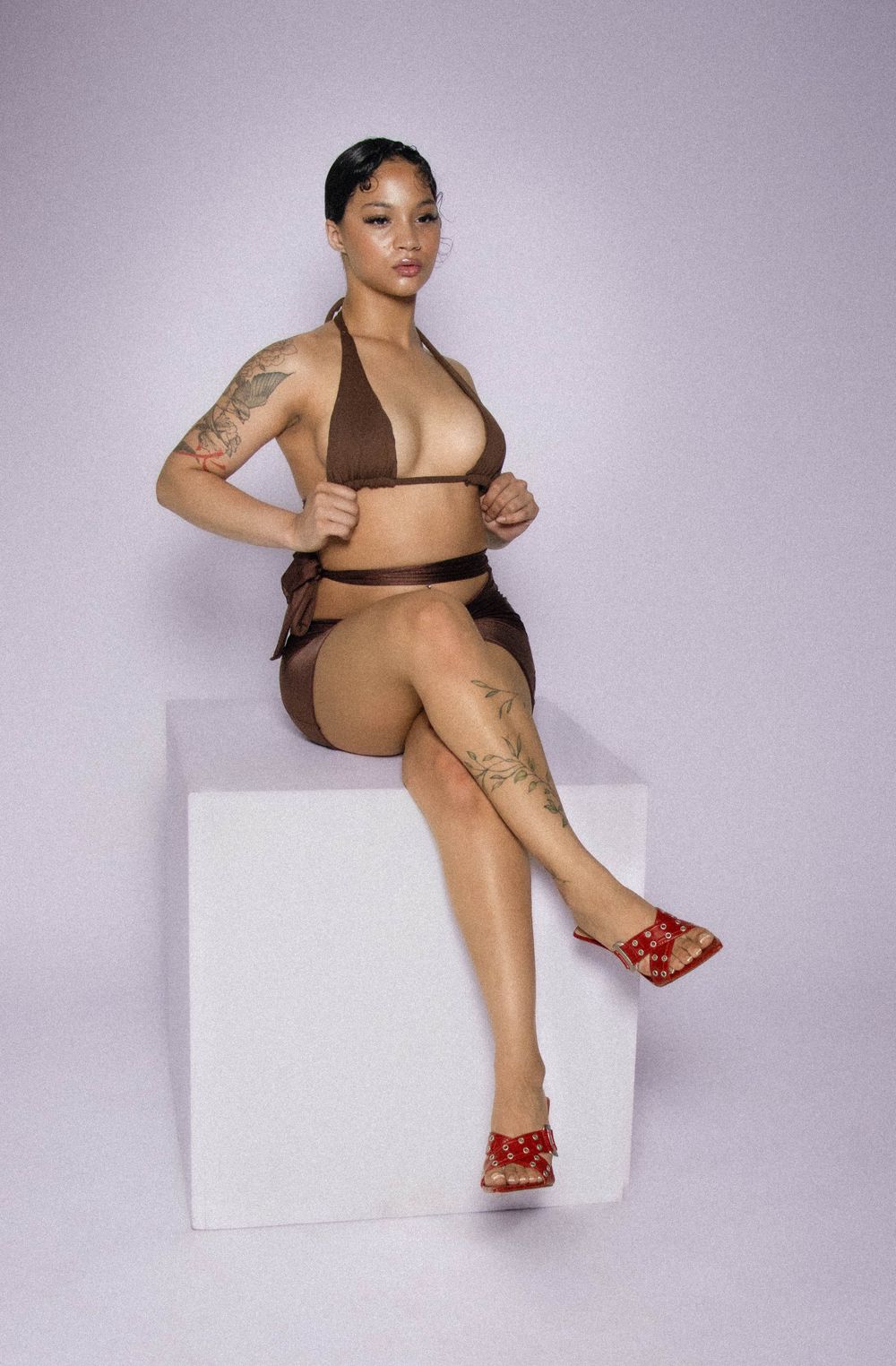 Model wearing brown two-piece swimsuit and red sandals poses on white cube against purple background.