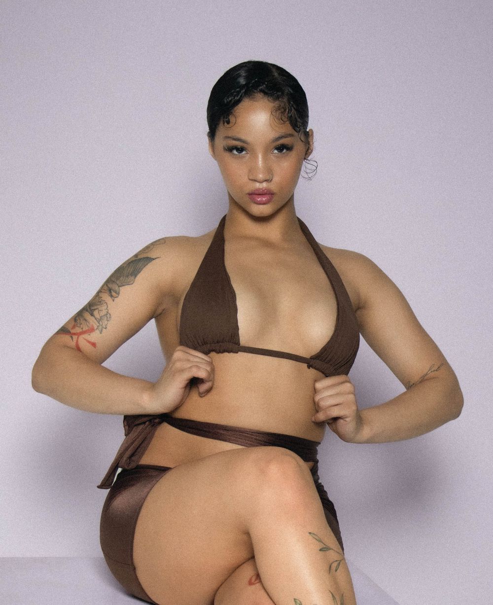 Model wearing a brown halter bikini swimsuit poses against purple backdrop showing visible tattoos on arm and thigh.
