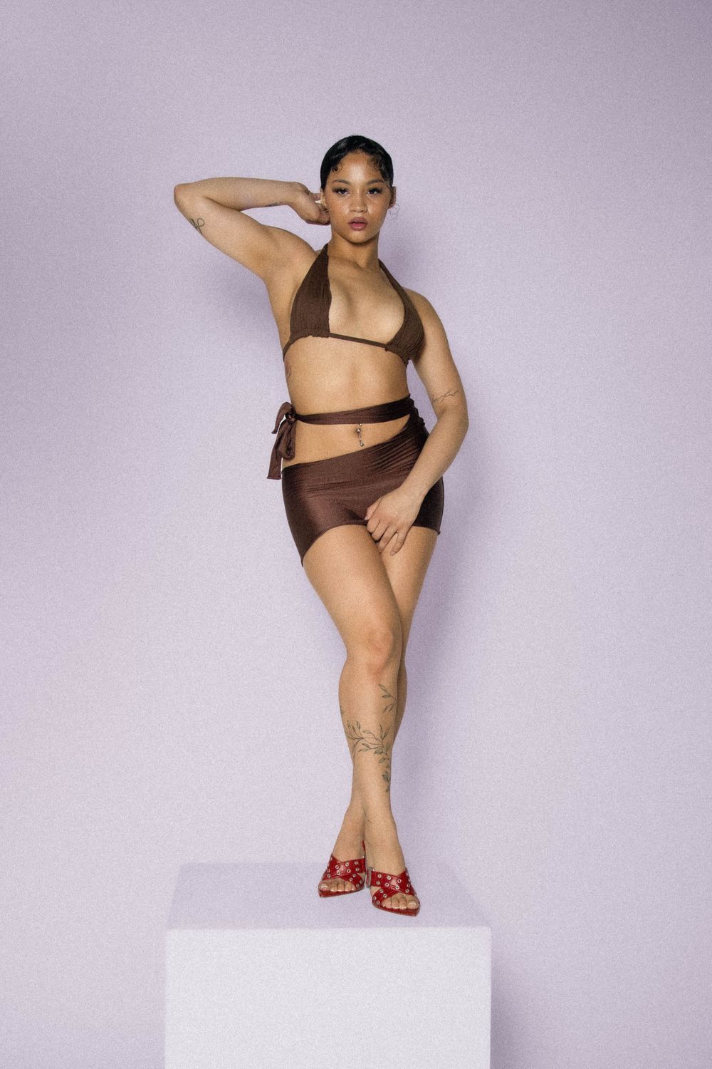 A model poses in a brown bikini and red sandals on a white pedestal against a lavender background.