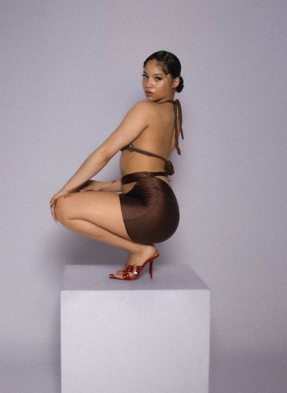 Model in brown outfit poses elegantly on white cube wearing red heels against gray studio backdrop.
