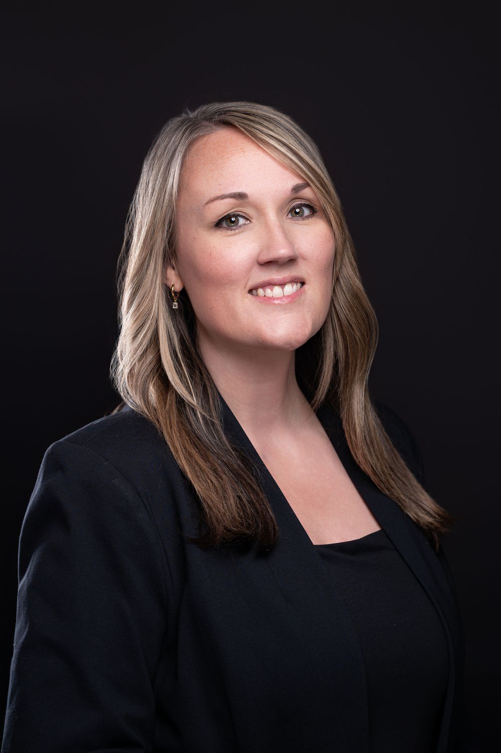 Professional headshot of someone wearing a black blazer and top against a dark background, smiling warmly.