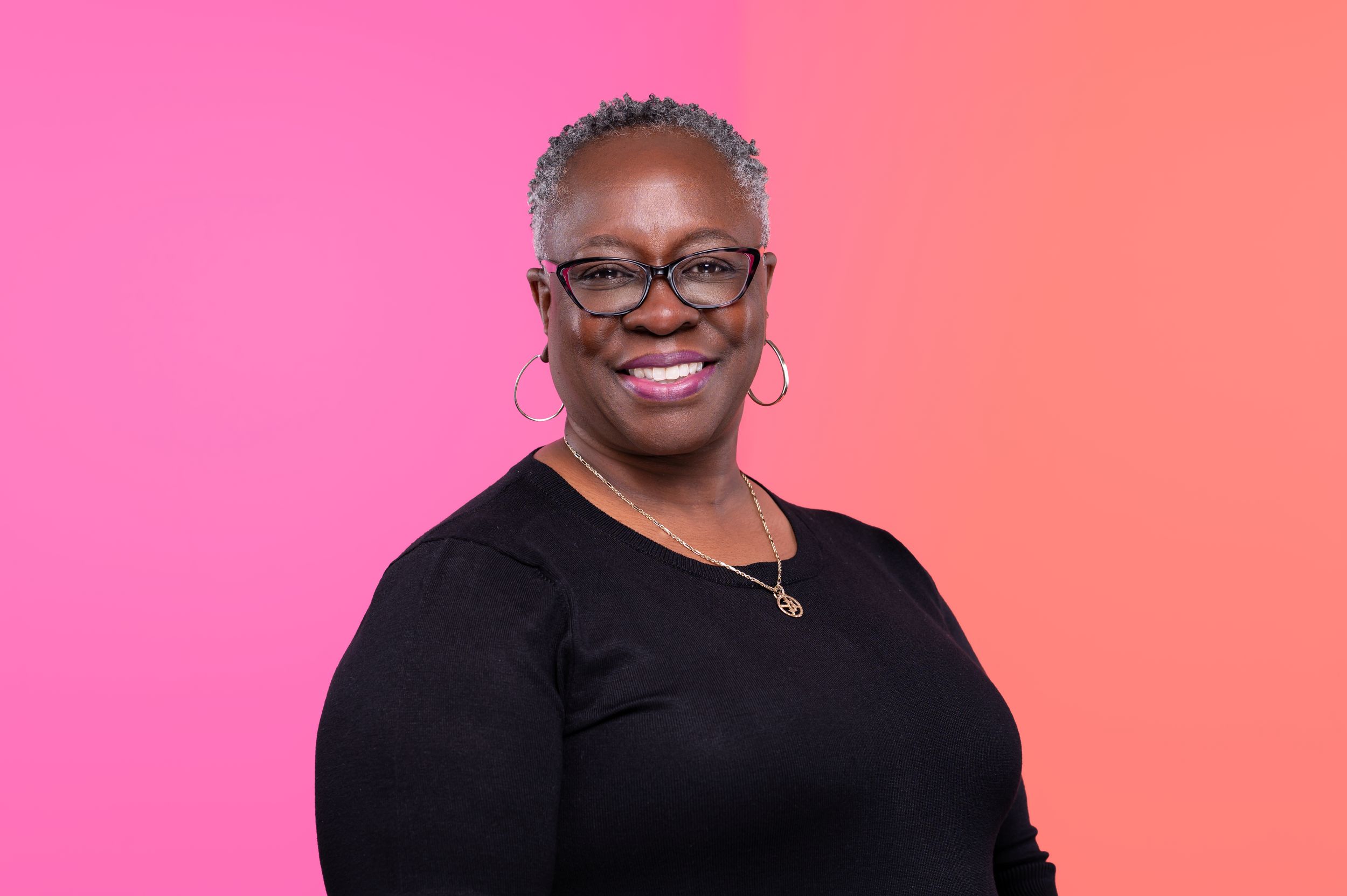 A person wearing glasses and a black top smiles warmly against a pink and orange gradient background.