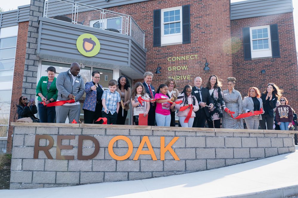 A group gathers for a celebratory ribbon cutting ceremony in front of a brick building with Red Oak signage.
