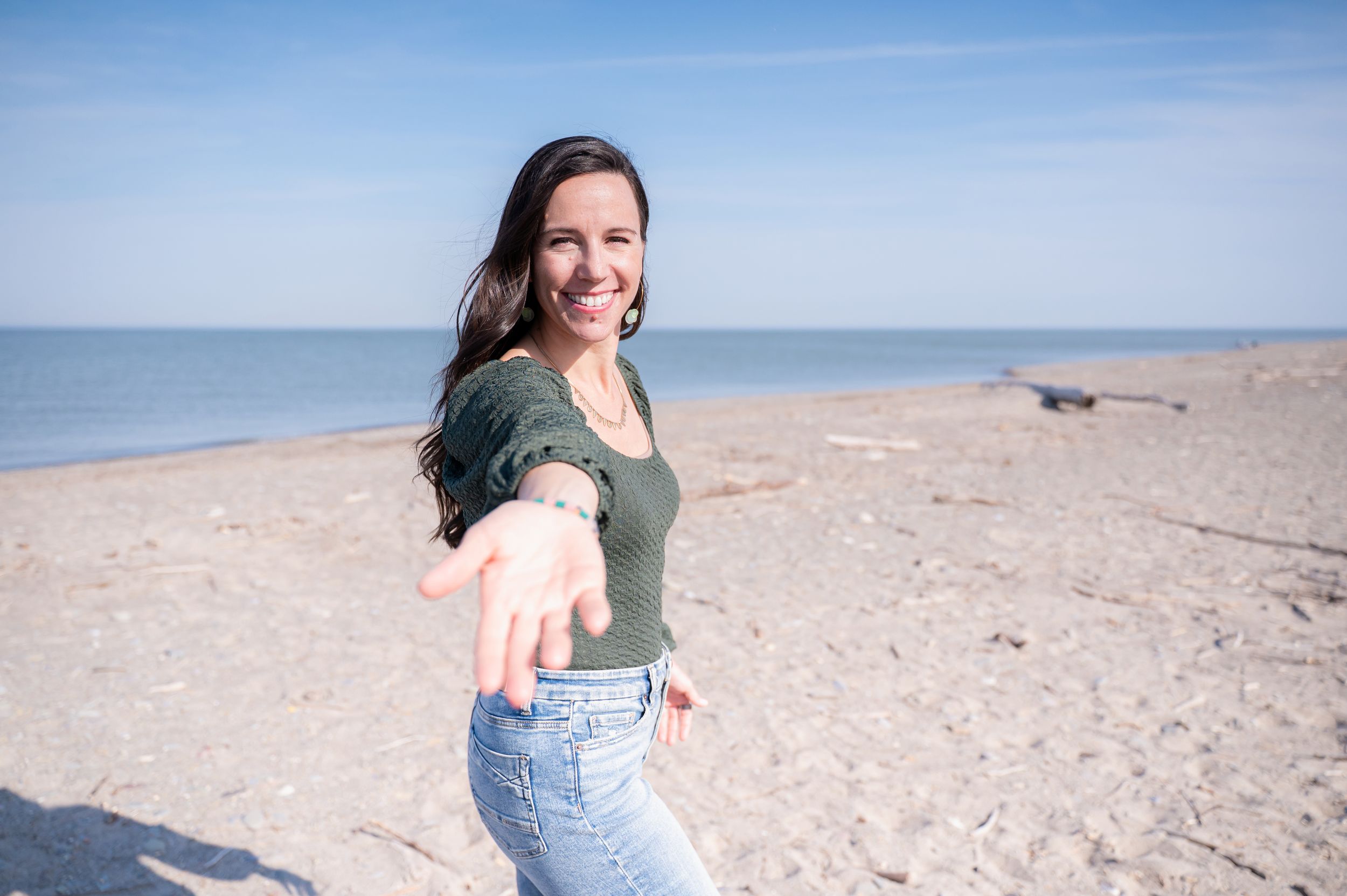 A person in casual attire reaches out towards the camera while standing on a sunny beach with ocean in the background.
