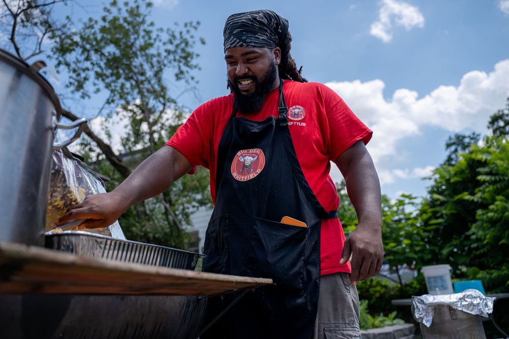 Smiling person wearing a red t-shirt and black apron while working at an outdoor cooking station.
