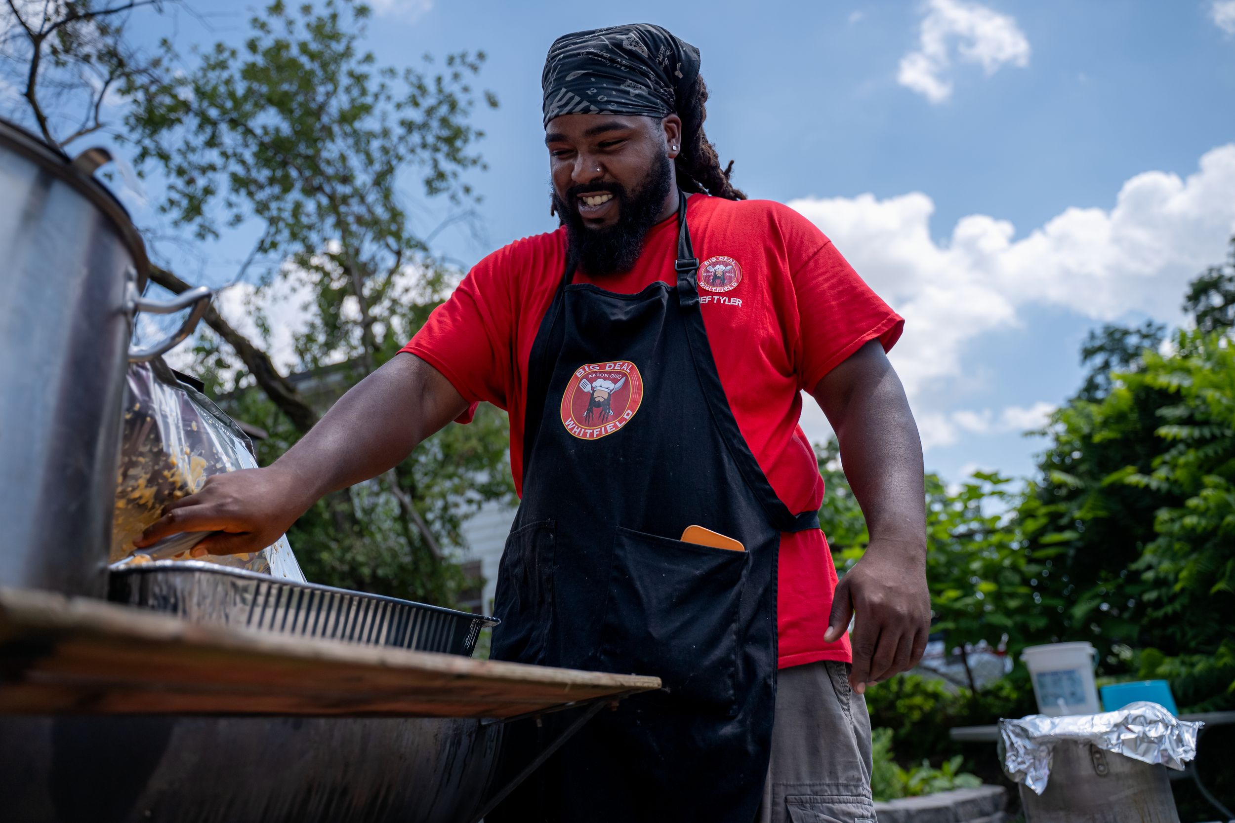 Smiling person wearing a red t-shirt and black apron while working at an outdoor cooking station.