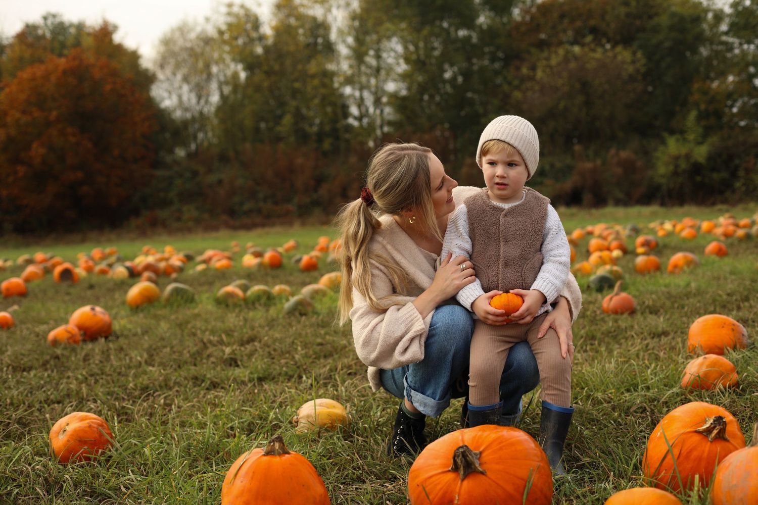 Two people sitting together in autumn pumpkin patch field surrounded by orange pumpkins during harvest season.