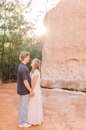 Couple in casual attire standing together against a large sandstone rock formation at sunset.