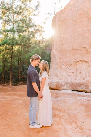 Two people holding hands while standing on red clay ground next to towering rock wall.