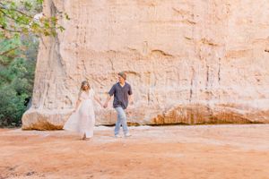 Couple walking hand in hand along a red clay path beside a massive rock formation.
