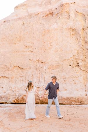Two people in casual dress holding hands near a tall sandstone cliff face.