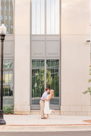 A couple embraces against a modern building exterior during their engagement session.