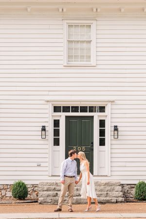 Couple stands in front of white colonial house with green door and classic architectural details.