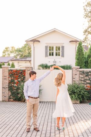 A couple dances on a brick patio in front of a white house with red flowers.