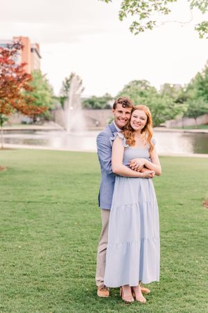A loving couple poses for a portrait with a fountain and green lawn in the background.