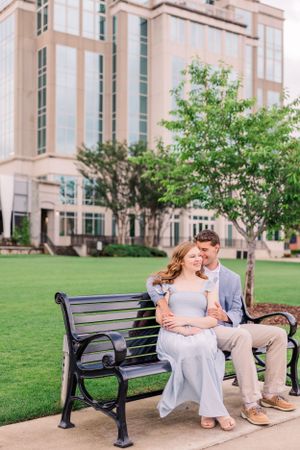 A romantic couple sits together on a park bench in front of a modern building.