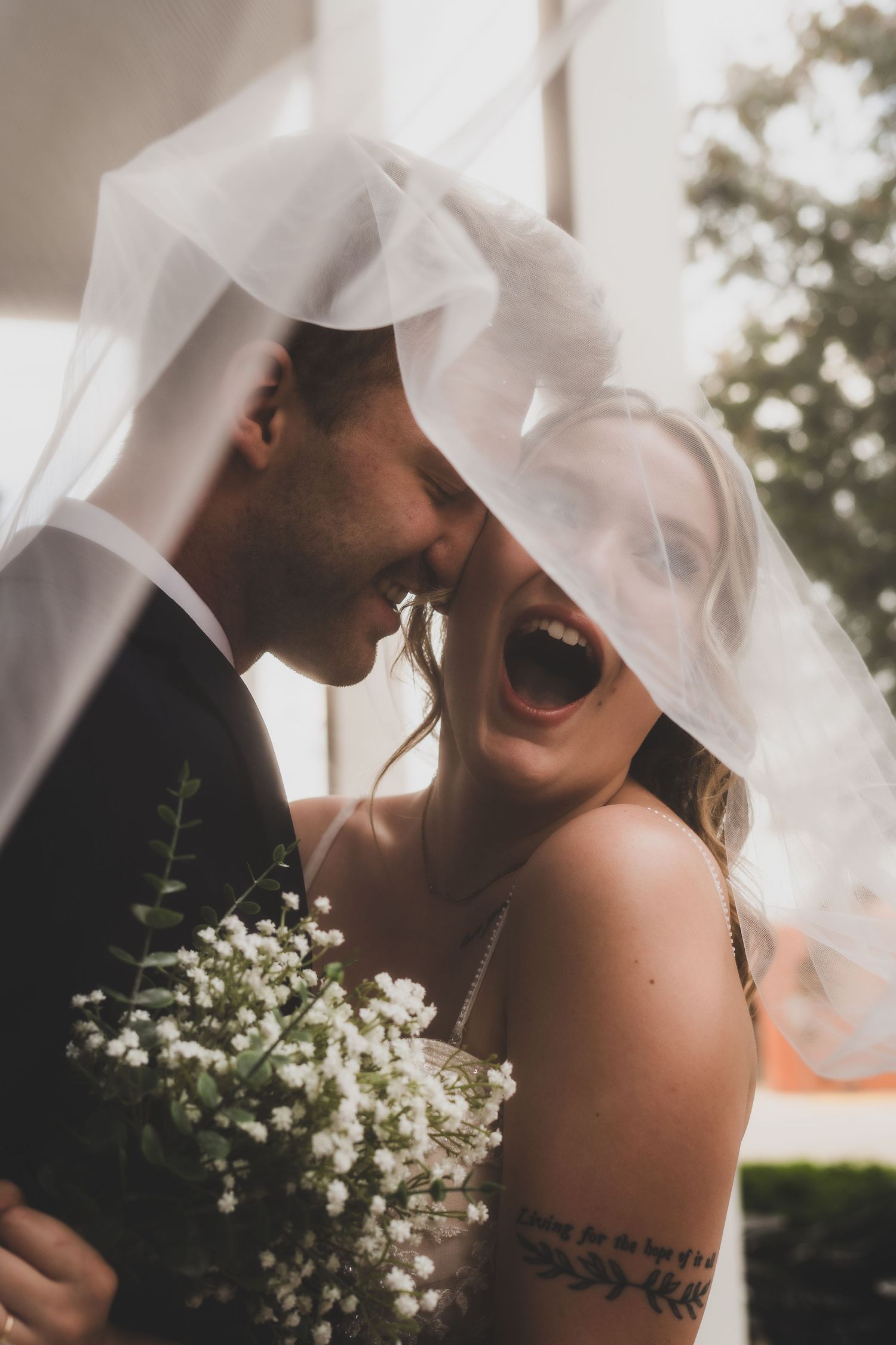 Newlywed couple embraces intimately under white columns with bride holding baby's breath bouquet and flowing veil.