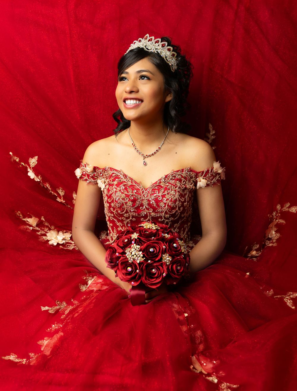 Traditional quinceañera portrait in red ball gown with crystal tiara against red backdrop.