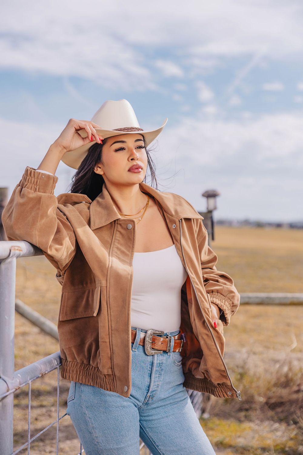 Woman in brown leather jacket, white tank, jeans, and cowboy hat leans on ranch fence outdoors.