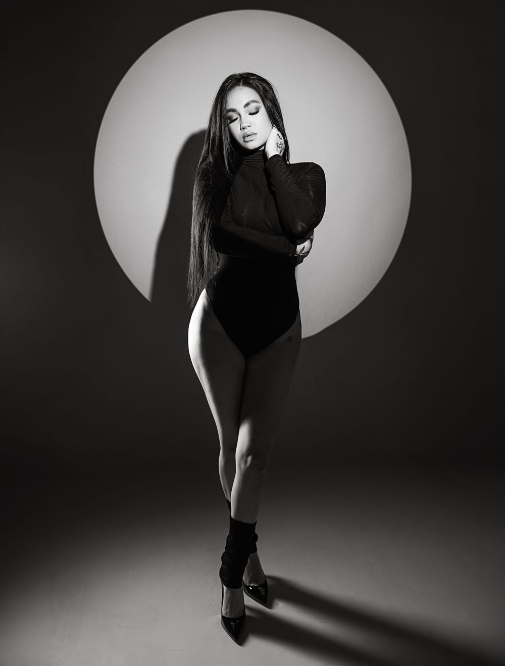 Black and white photo of a woman in a black bodysuit and heels posing against a circular light backdrop.