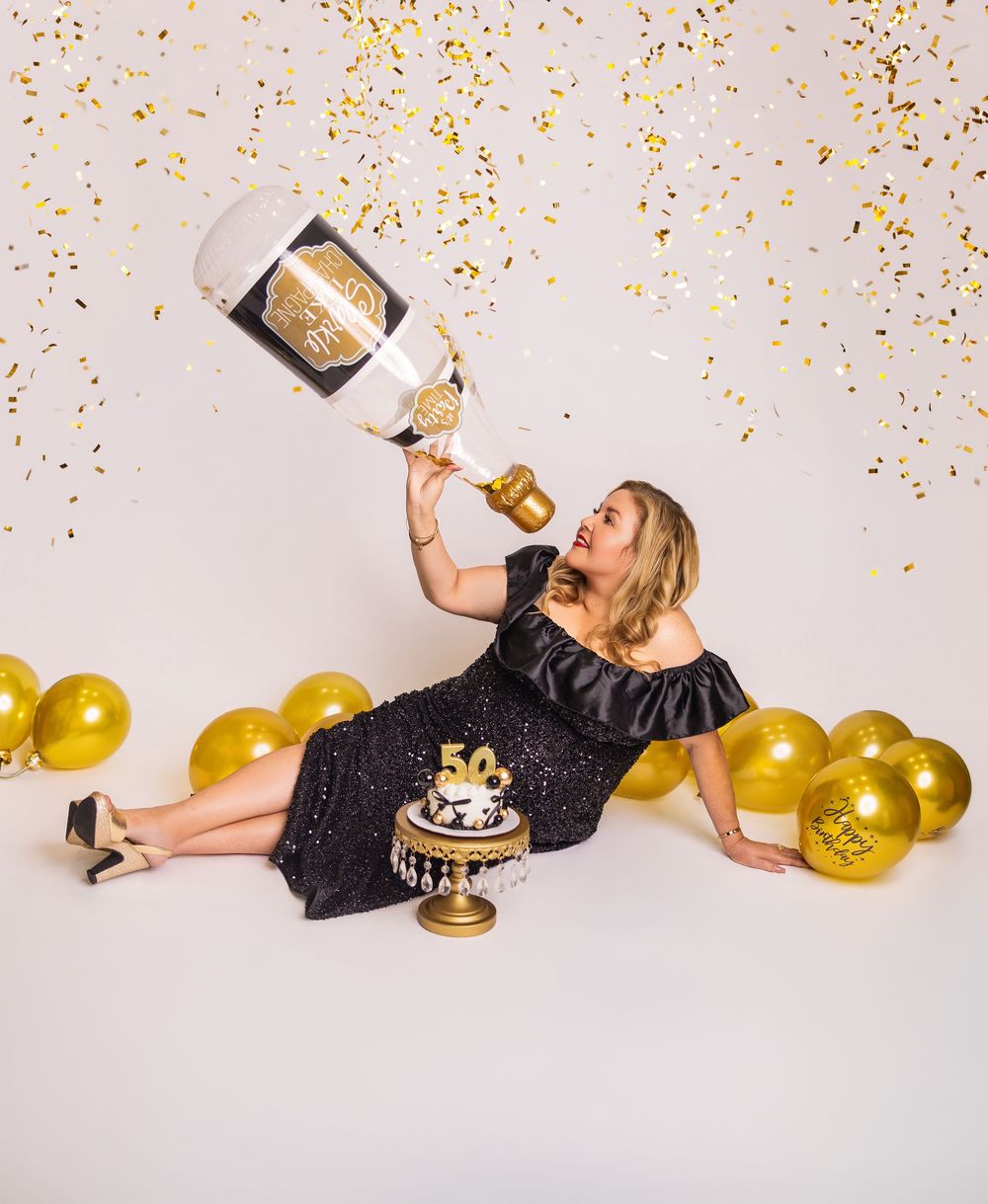 Woman in black sequin dress celebrating 50th birthday with giant champagne bottle, gold balloons, confetti, and cake.