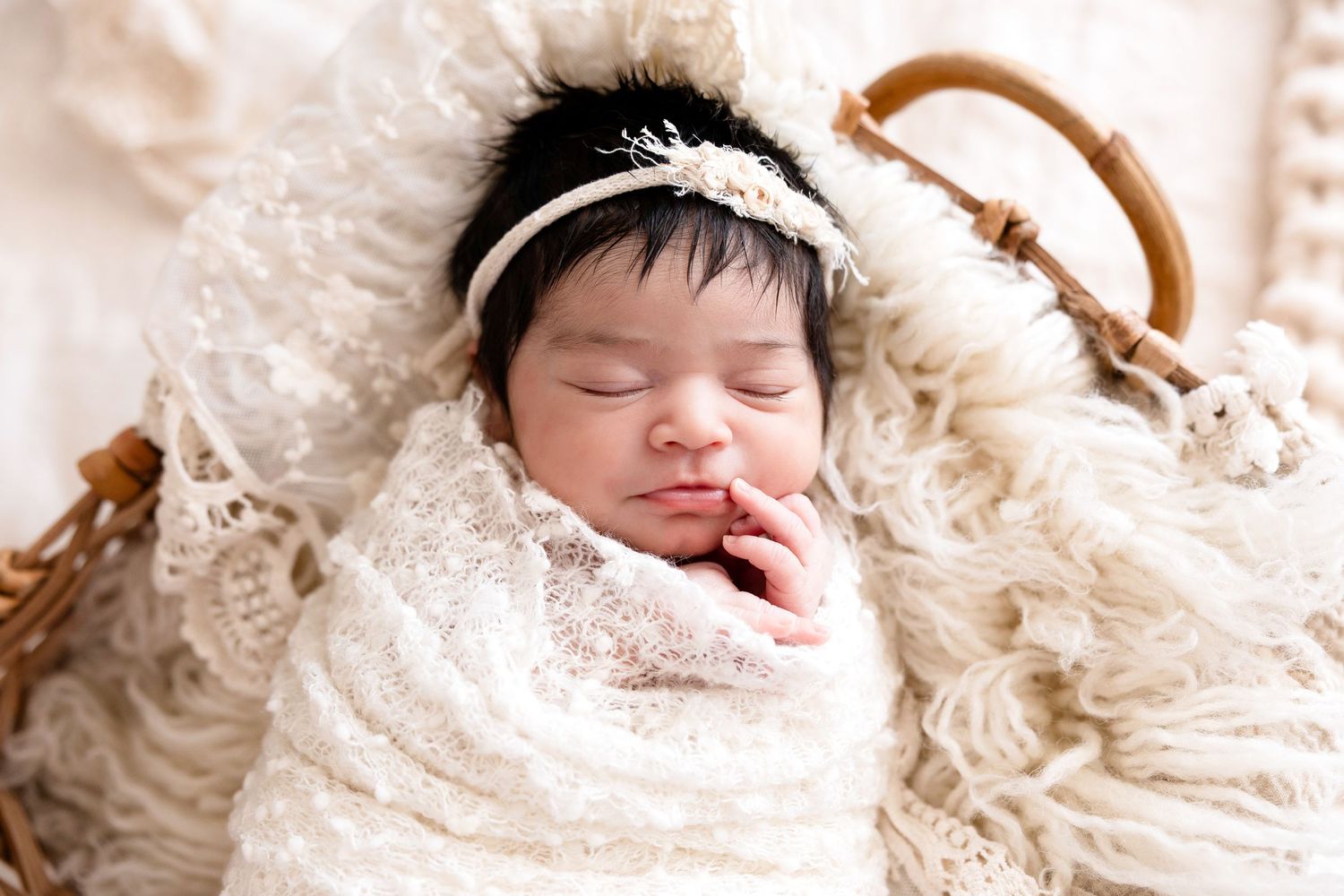 Sleeping newborn baby wrapped in white lace swaddle with floral headband resting in a wicker basket.