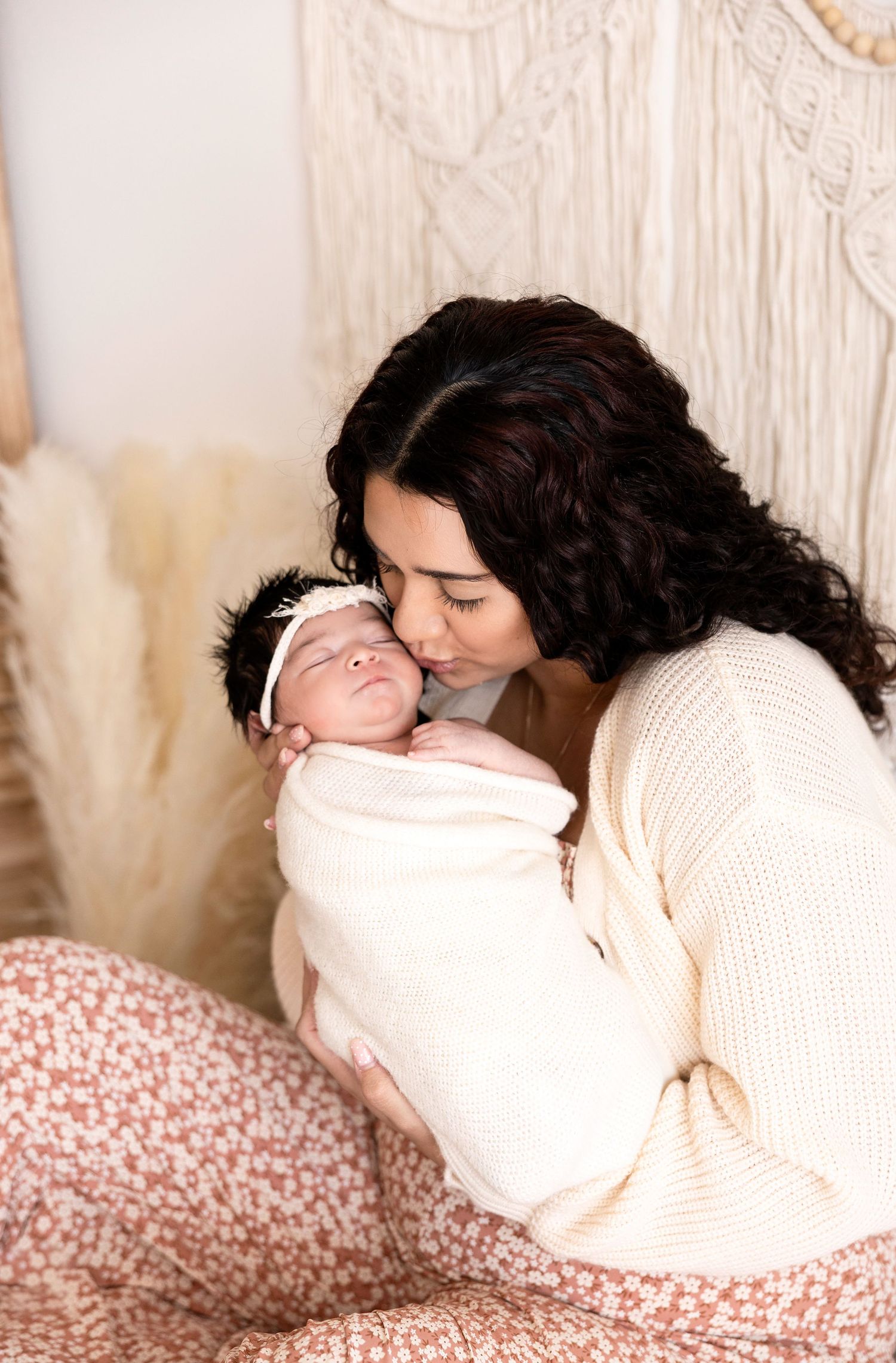 A mother with dark curly hair gently kisses her swaddled newborn baby wearing a white headband.