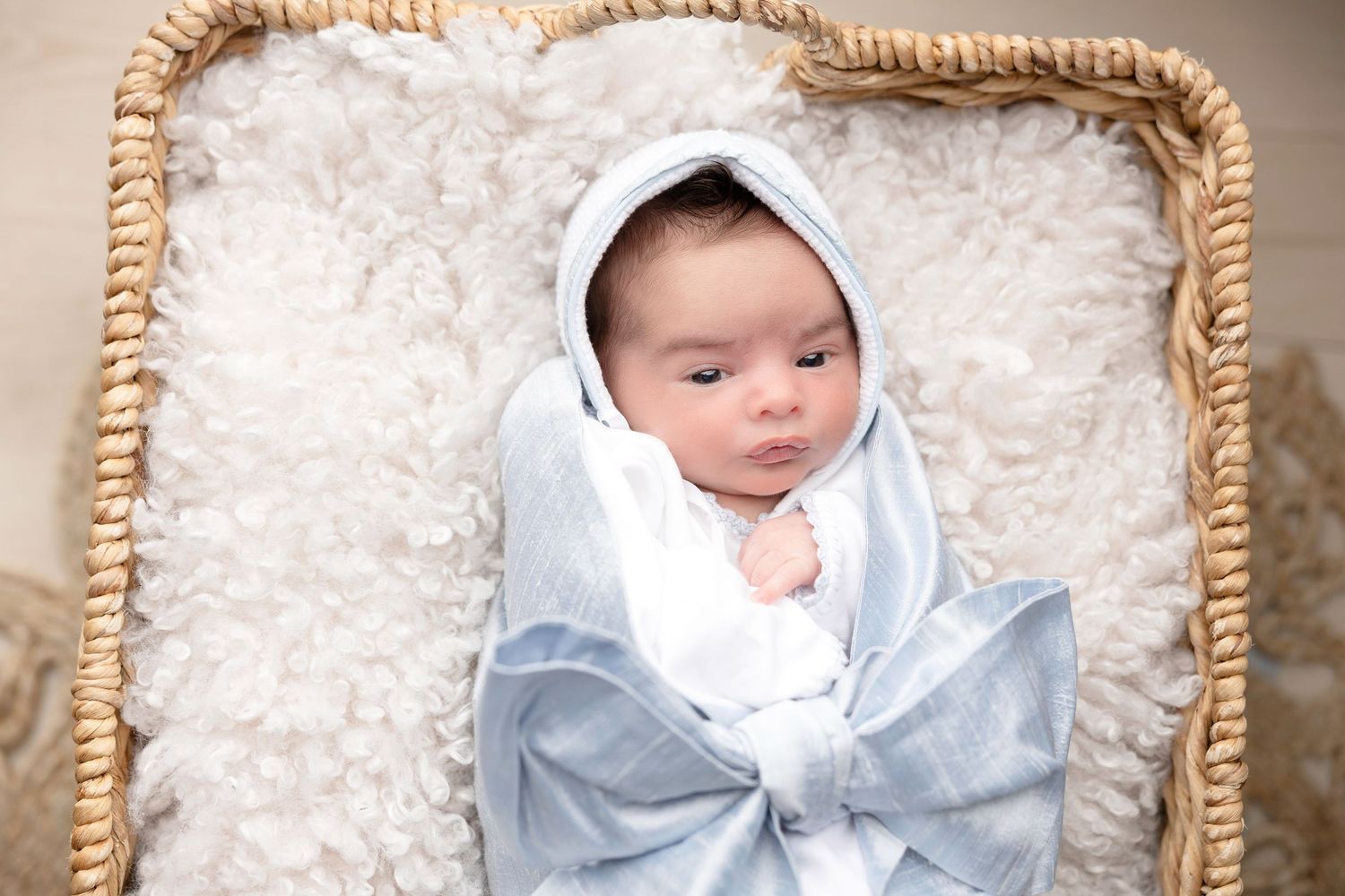 Newborn baby resting in a woven basket with soft white bedding and blue blanket.