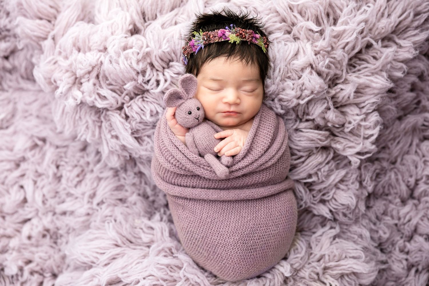 Newborn baby girl wrapped in purple knit swaddle, holding tiny stuffed animal, wearing floral headband on fluffy lavender rug.